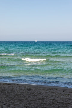 Calm turquoise waves roll onto a sandy beach under a clear blue sky, with a distant sailboat on the horizon, evoking peace and relaxation.