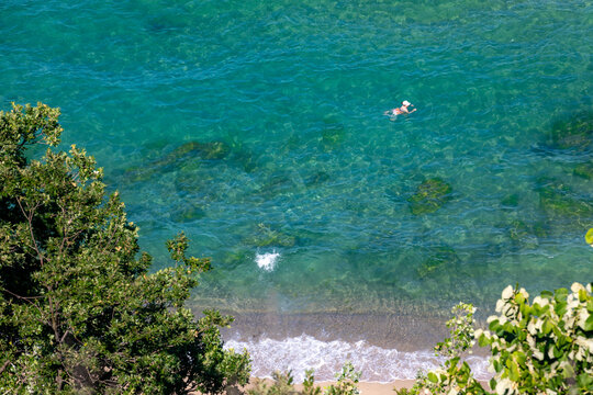 A person swims in transparent turquoise water near a sandy shoreline, surrounded by lush green trees, captured from above on a sunny day.