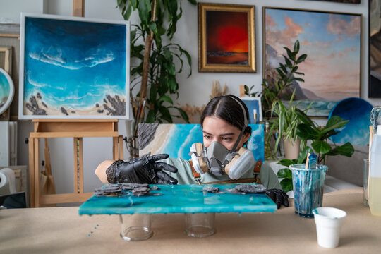 Talented focused craftswoman fully engaged in working process on epoxy resin art in workshop, female artist carefully adjusting edge of painting with hand in protective glove before resin hardens.
