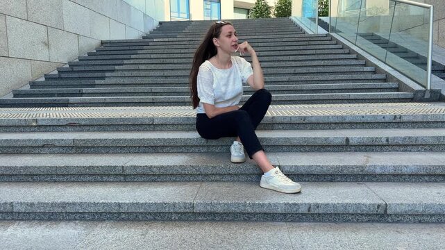 Young woman sitting on wide outdoor steps while talking on a smartphone, urban lifestyle, communication, casual city break.