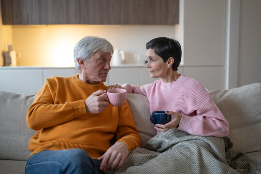 Senior husband and wife sitting on couch at home together drinking tea and talking with each other, elderly partners discussing and expressing emotions, comfort and trust in family relationships.