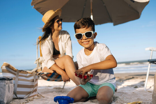 Mother and Son Playing on Sandy Beach Under Umbrella During Sunny Summer Vacation Together