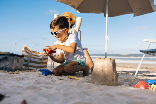 A mother relaxes in a beach chair under an umbrella as her young son concentrates on building a sandcastle nearby. Warm sunlight, seaside breeze and family vacation vibes create a peaceful scene.