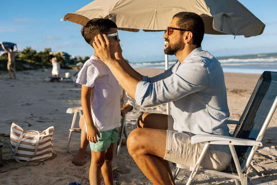 A father gently puts sunglasses on his young son beneath a beach umbrella, sharing a warm moment of parental care and sun protection during a relaxed summer family vacation by the ocean.
