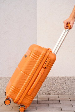 Person pulls an orange rolling suitcase along a paved walkway beside a light wall