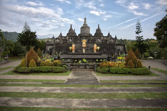 Magnificent mini replica of Borobudur temple surrounded by lush gardens, pathways, and golden Buddha statues under a blue sky at Brahma Vihara Arama monastery in Bali.