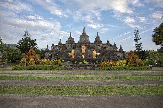 Magnificent mini replica of Borobudur temple surrounded by lush gardens, pathways, and golden Buddha statues under a blue sky at Brahma Vihara Arama monastery in Bali.