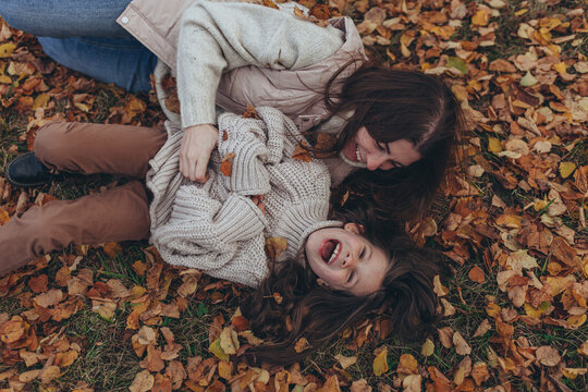 Happy mother and daughter laughing and playing together in park covered with autumn leaves. Mother's day, family bonding, unconditional love, joy of childhood, happiness and seasonal lifestyle.