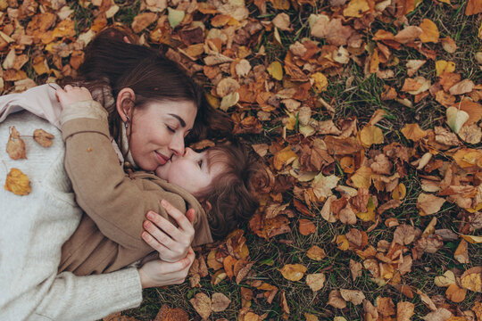 Loving mother embracing  child while lying together on bed of fallen autumn leaves in park. Unconditional love, maternal care, parenting, emotional connection, autumn lifestyle, family happiness.