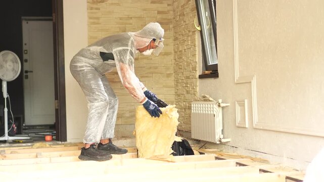 A cinematic medium shot of a worker wearing a hazmat protective suit and a respirator, packing fiberglass insulation into a black garbage bag during floor repair.