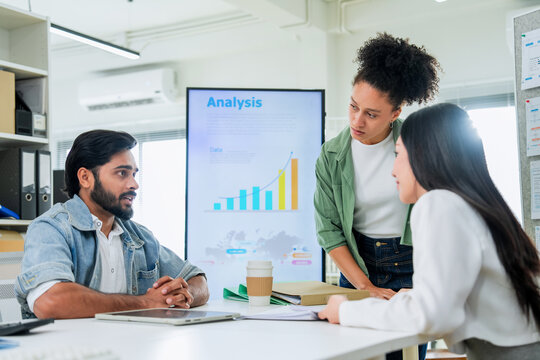 A woman points to data on a tablet while explaining analysis results to two colleagues, a bar chart displayed on the screen behind them a focused scene of business data review and team analysis