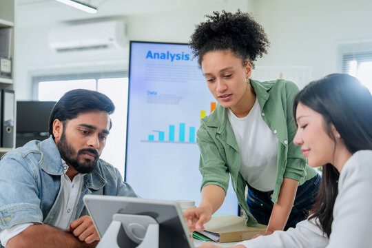 A hand reaches to point at a tablet screen amid folders and documents, colleagues leaning in to follow along a close, process-driven moment of collaborative problem-solving at work