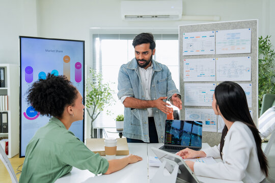 A man presents market share data on a large screen while explaining to two colleagues at a desk, one reviewing stock charts on a laptop a comprehensive scene of business analysis and team presentation