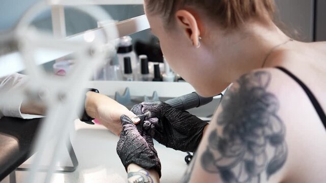 Nail technician removes old nail polish from a client's fingernail using an electric nail drill. Professional manicure, nail care and beauty salon service concept