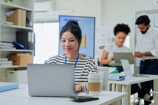 A focused employee works on her laptop in the foreground while colleagues collaborate at a desk behind her, capturing the layered activity of a busy, modern open-plan office in full swing.