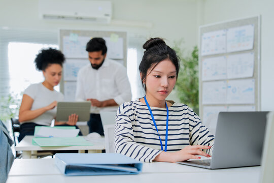 A focused employee works on her laptop in the foreground while colleagues collaborate at a desk behind her, capturing the layered activity of a busy, modern open-plan office in full swing.
