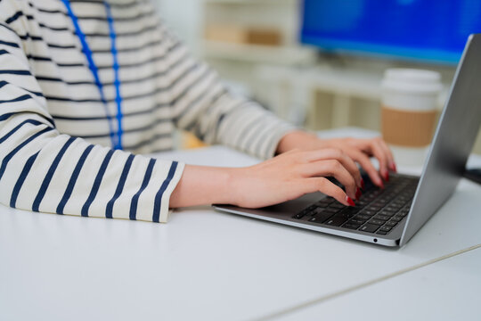 Closeup of hands with red-painted nails typing on a laptop keyboard, a blue lanyard and coffee cup visible in the blurred background a faceless, focused image of professional office work.