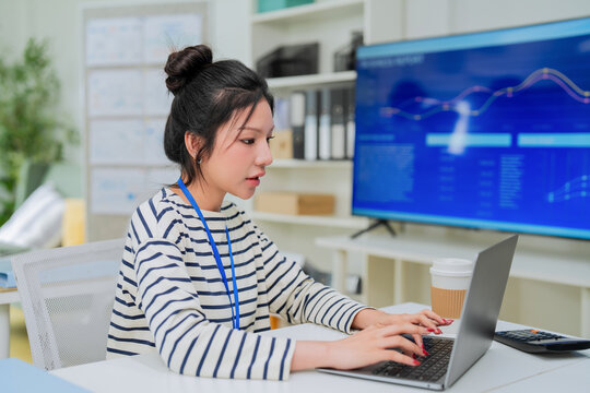 A focused employee wearing a lanyard types on her laptop, a large monitor behind her displaying colorful performance analytics charts a sharp depiction of data analysis and business intelligence work.