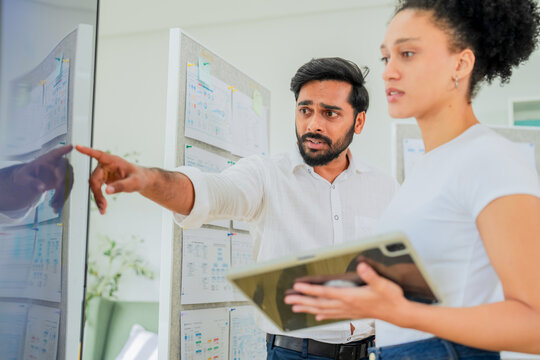 A man points intently at a data board while explaining details to a colleague holding a tablet, both focused on analyzing project information a dynamic scene of workplace analysis and strategic discus