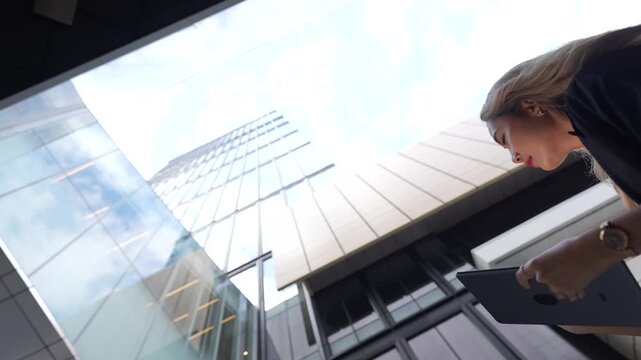 Asian businesswoman crouches slightly while writing on her tablet, the glass skyscraper stretching upward behind her in a striking extreme low angle shot in the business district.