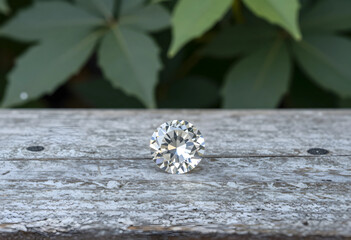 Obraz premium Brilliant cut diamond on weathered wooden surface with green leaves in soft focus background, the faceted gemstone reflects light, its clarity and sparkle
