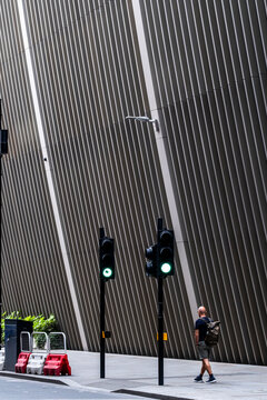 Beside a ribbed building facade, a pedestrian crosses open pavement as a green traffic signal marks a street in London, England, beneath stark urban architecture.