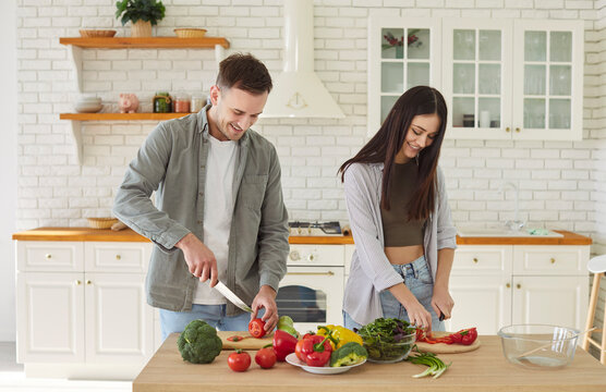 Happy young family couple, man, woman cutting salad, helping, healthy vegans cooking at home in modern kitchen, vegetarian diet, vegetables, plate of fresh eco food at table, health benefits