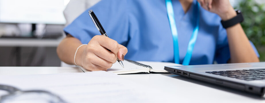 Healthcare worker scrubs pen notebook laptop hand desk writing medical office blue healthcare worker scrubs writing notes with pen notebook beside laptop desk, focused professional recordkeeping