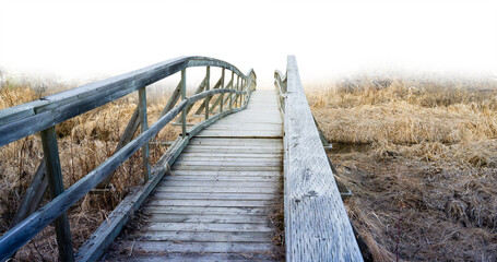 Obraz premium View over an old dilapidated wooden walking bridge surrounded by dry brown grass with a transparent sky 