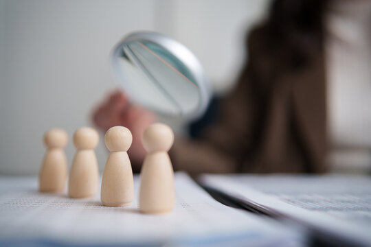 Recruiter examining wooden candidate figures with a magnifying glass, representing recruitment, applicant screening, talent search, employee evaluation, hiring decisions and human resources.
