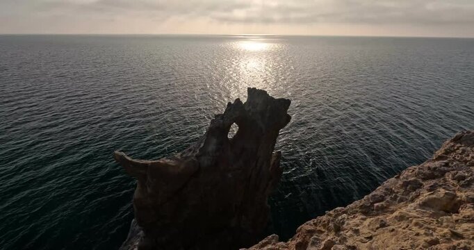 Sunlight glistens on the open sea beside a perforated monolithic rock tower along the coastal cliff. High-angle static shot.