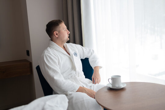 Side view profile of a relaxed millennial male holding a cup of tea while sitting in an armchair near a large window. Concept of morning routine, relaxation, luxury spa wellness, and hospitality