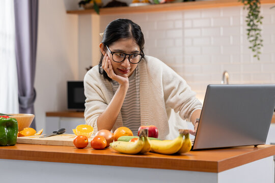 Woman laptop kitchen fruit eyeglass home table casual Asian technology, focused woman using laptop in kitchen with fruit on table, home interior for remote work, online study, healthy lifestyle