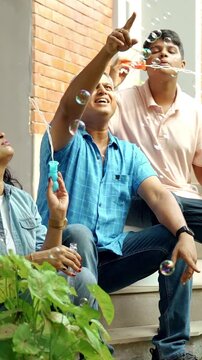 Indian Asian family enjoying quality time outdoors, mother and son making soap bubbles while the father watches and the daughter playfully tries to burst them, all sitting together on stairs of home