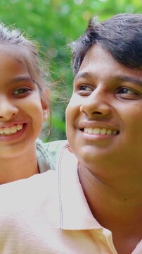 Indian Asian big brother joyfully carrying his small sister on his back, giving her a playful piggyback ride outdoors, bonding in nature on a sunny day