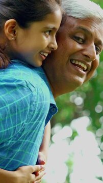 Indian Asian father with gray hair in his forties carrying his daughter on his back, giving her a joyful piggyback ride outdoors, bonding in nature on a sunny day