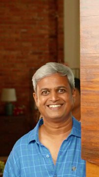 Indian Asian family of four joyfully welcomes guests to their home, as parents in their forties and their teenage son and small daughter open the big wooden entrance door with warm smiles