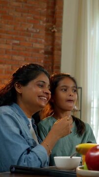 An Indian Asian family of four, with parents in their forties and two kids, enjoying a meal together at the dining table. The mother serves food while they talk and share happy moments
