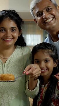 Indian Asian mother joyfully fries a dish cooking food in the kitchen, surrounded by her excited husband and two kids, all eagerly watching her culinary skills