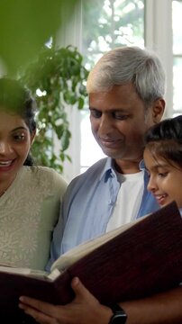 A closeup of a happy Indian Asian family consisting of parents in their mid-forties, a teenage son, and a small daughter joyfully reminiscing together while flipping through a cherished photo album