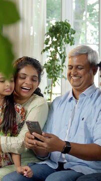 Indian Asian family of four, with parents in their forties and two kids, sitting closely on a sofa laughing out loud while enjoying a funny video on a smartphone, sharing a joyful moment together