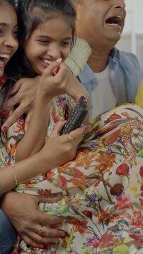 Indian Asian family of four, parents in their forties and two kids, sitting closely on a sofa, laughing out loud while watching television together, enjoying a fun-filled family evening at home