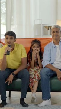 Indian Asian family of four, parents in their mid forties, a teenage son, and a small daughter celebrate a sports victory while watching TV, waving the Indian tricolor flag with excitement.