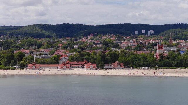 Aerial video captures the sandy Sopot beach, calm sea water, historic buildings, and town in Poland under a cloudy sky with people relaxing near the coast.