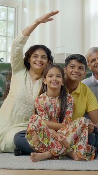 In this close up photo, an Indian Asian family of four,parents in their mid forties, a teenage son, and a small daughter,sit on a cozy carpet, smiling joyfully, creating a roof sign with their hand