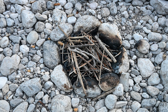 Small rustic campfire pit built with river rocks on a pebble bank. Dry twigs and branches arranged inside a stone fire ring for camping.