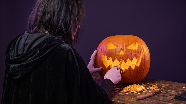 Woman in black cloak carving a glowing pumpkin for Halloween. Person making jack-o-lantern for autumn holiday decoration.