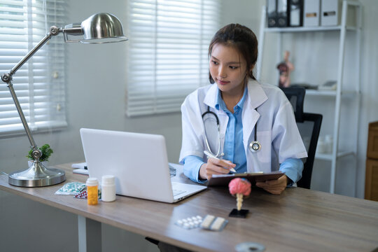 Young Asian female doctor writing notes on clipboard while consulting laptop in modern medical office