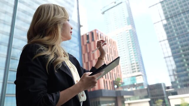 Businesswoman in side profile raises a stylus while holding her tablet against a backdrop of glass towers, gesturing as if sketching an idea in the open air district.