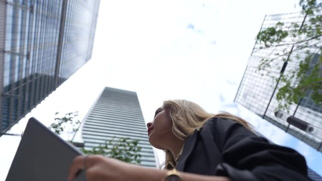 Businesswoman holds a tablet at a dutch angle beneath leaning glass skyscrapers, tilting her chin upward as the static tilted camera exaggerates the towering perspective.